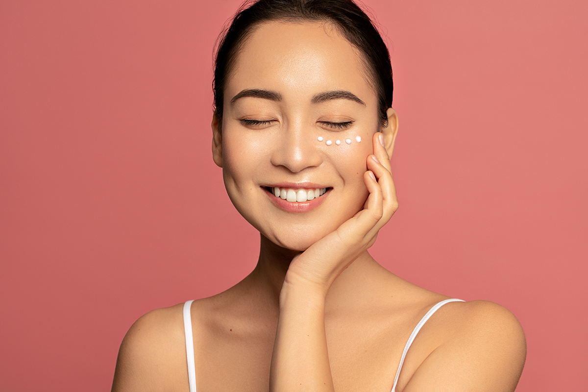 Young beautiful model posing against pink background with applied cream under her eyes